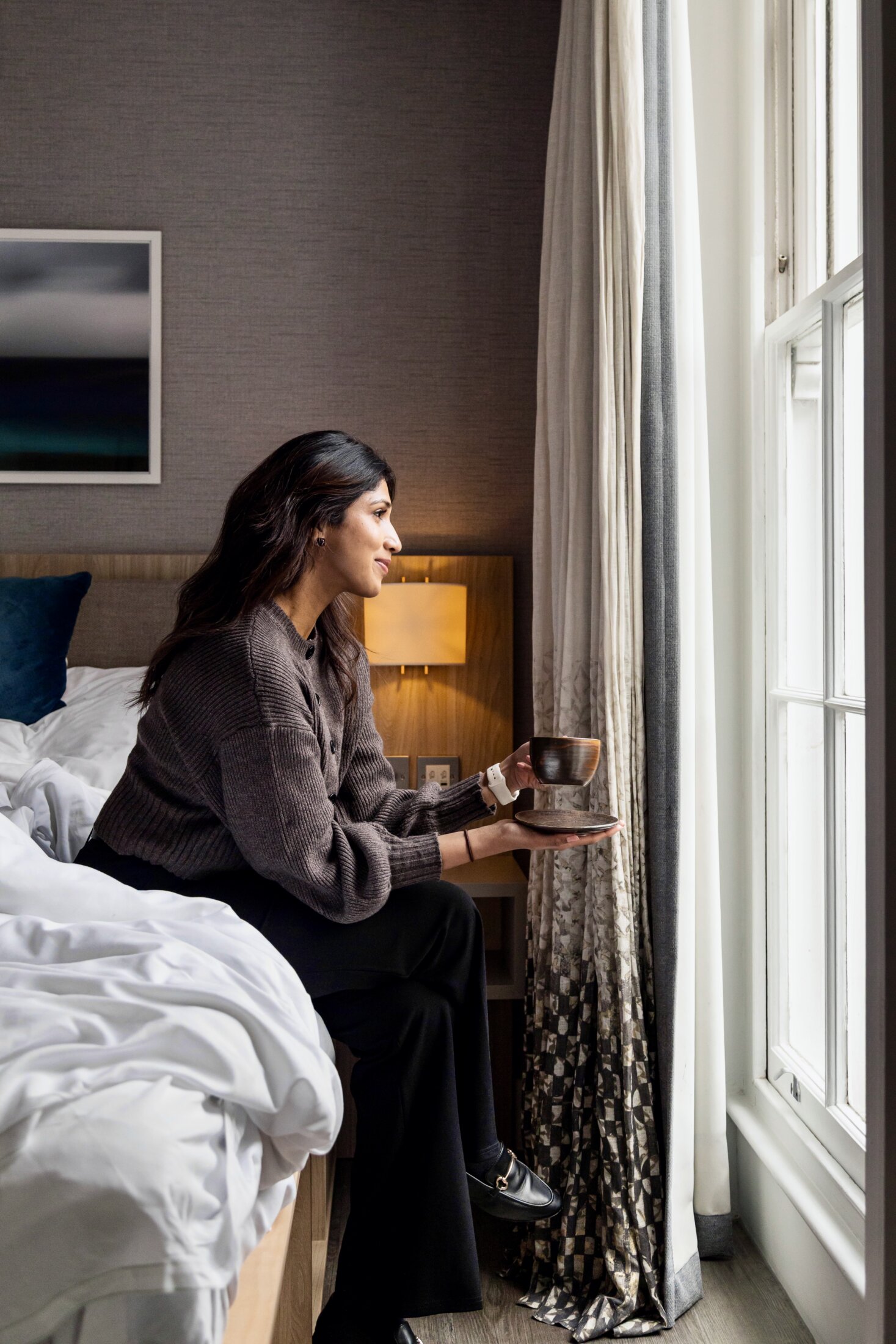 Guest sitting on the bed looking out of the window with a cup of coffee at Inhabit Queen's Gardens hotel